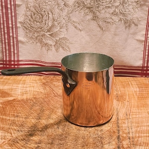 Peut inclure: Une casserole cylindrique en cuivre avec une poignée en métal foncé fixée par deux rivets. La surface extérieure est en cuivre poli, tandis que l'intérieur est en métal argenté. Un petit tampon circulaire est visible sur le côté du corps en cuivre.