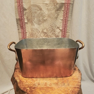 May include: A copper-colored rectangular tub with curved handles. The interior is a lighter metal. The tub sits on a wooden surface, with a rooster-themed fabric in the background. The tub is likely used for decorative or functional purposes.