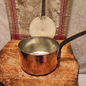 May include: A copper saucepan with a black handle sits on a wooden surface. A round, metal lid and a long-handled metal spatula rest behind the pan. The copper pot has a shiny, reflective surface, and the handle is riveted to the side.
