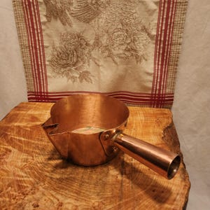 May include: A copper-colored saucepan with a long handle and a spout. The pan is sitting on a wooden surface. The background features a floral patterned fabric with red stripes.