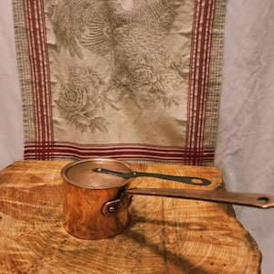 May include: A small copper saucepan with a lid and a long handle. The pan sits on a wooden surface, with a decorative fabric in the background. The fabric has a floral and bird design with red stripes.