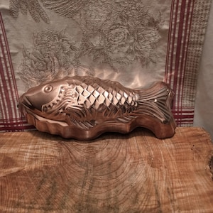 May include: A metallic copper-colored fish-shaped mold featuring detailed embossed scales, fins, and a defined eye. The mold has a shiny, reflective surface and is shaped like a swimming fish with a textured tail and head design.