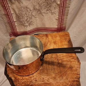 Puede incluir: Una cacerola de cobre con un asa negra descansa sobre una superficie de madera. El interior de la olla es plateado. Un tejido con estampado floral y rayas rojas está en el fondo.