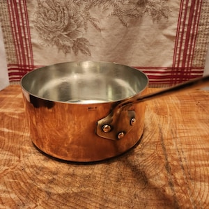 May include: A copper saucepan with a long handle and a shiny interior. The pot has a rounded shape and is attached to the handle with rivets. The words "Made in France" are stamped on the side of the pot.
