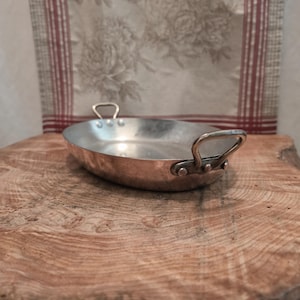 Pode incluir: Uma travessa de servir oval em cobre com revestimento interior metálico. A peça possui duas pegas metálicas curvas nos lados opostos, cada uma fixada firmemente ao corpo de cobre com três rebites visíveis para um manuseamento seguro.