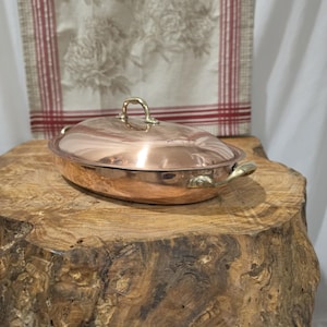 Puede incluir: Una fuente de asar ovalada de cobre con tapa a juego y dos asas laterales de latón. La tapa cuenta con un asa central de latón. La superficie de cobre pulido refleja la luz, destacando el acabado metálico y la forma redondeada del objeto.