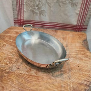 Puede incluir: Una fuente de servir ovalada de cobre con un revestimiento interior plateado pulido. El recipiente cuenta con dos pequeñas asas metálicas curvas en los lados opuestos y el número 26 está grabado claramente en el borde de cobre cerca de un asa.