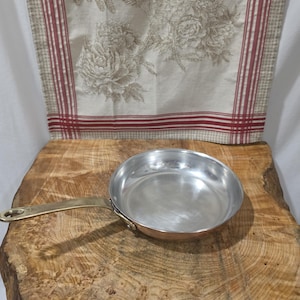 Può includere: Una padella rotonda in rame con rivestimento interno in acciaio inossidabile lucido e un lungo manico curvo in ottone fissato con due rivetti. La padella presenta una finitura metallica liscia e una base piatta per la cottura sui fornelli.