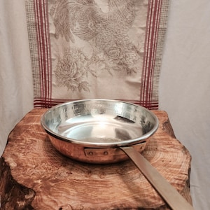 May include: A copper-bottomed frying pan with a long, flat handle. The pan is set on a rustic, wooden surface. A decorative fabric with a rooster and floral design hangs in the background.