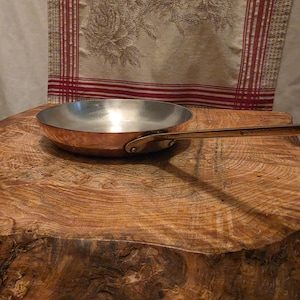May include: A round copper frying pan with a polished silver-toned interior and a long, slender metal handle attached by two rivets. The exterior of the pan features a warm, metallic copper finish, while the interior surface is smooth and reflective.