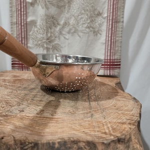 May include: A copper colander with a polished stainless steel interior and a long wooden handle. The bowl features a series of small, evenly spaced drainage holes and is attached to the handle with a brass-colored metal bracket.