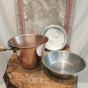 May include: A copper pot with a wooden handle, a matching metal lid, and a separate stainless steel colander insert. The pot has a wide, flared opening and a polished exterior finish, while the colander features a perforated base and sides.