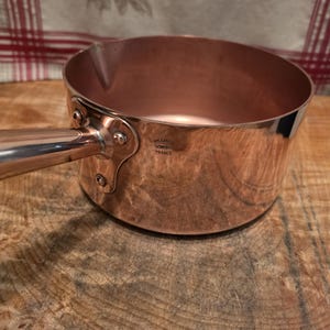 May include: A polished copper saucepan with a long, straight handle attached by a metal plate with three rivets. The side of the pan features a stamped mark that reads Williams Sonoma France. The interior is smooth and metallic in appearance.