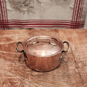 Może przedstawiać: Mały, miedziany rondel z pokrywką i dwoma zakrzywionymi uchwytami. Pokrywka ma mały, prostokątny uchwyt. Garnek stoi na drewnianej powierzchni. Tło obejmuje tkaninę we wzory kwiatowe z czerwonymi paskami.