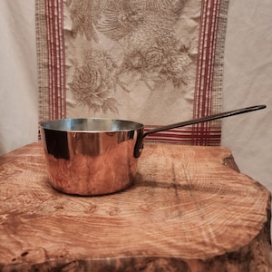 May include: A copper saucepan with a long, dark handle. The pot has a shiny, reflective surface and sits on a wooden surface. The background features a patterned fabric with floral and striped designs. This kitchenware is ideal for cooking.