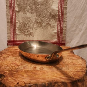 Puede incluir: Una sartén de color cobre con un mango largo. La sartén tiene un interior brillante y está colocada sobre una superficie de madera. El mango está sujeto con dos remaches. Un tejido decorativo con un diseño de gallo está en el fondo.