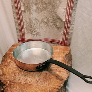 May include: A round copper sauté pan with a silver-toned interior and a long, black metal handle. The pan features a polished copper exterior and a sturdy, riveted handle attachment, showcasing a classic design for culinary use in a kitchen.