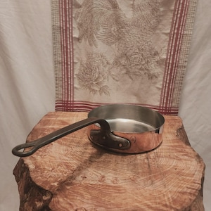 May include: A round copper skillet with a polished metallic interior and a long, dark, riveted iron handle. The exterior features a warm, reflective copper finish, while the handle is securely attached with three visible metal rivets.