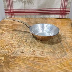 Puede incluir: Una sartén pequeña y redonda de metal martillado con un mango largo, delgado y ligeramente curvado. El interior de la sartén tiene un acabado plateado brillante y reflectante, mientras que el exterior y el mango tienen un tono cobrizo cálido.