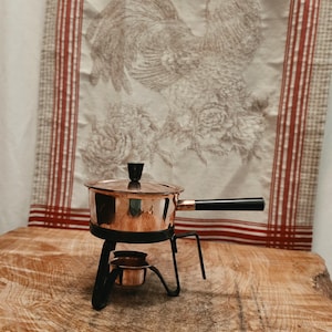 May include: A copper fondue pot with a black lid handle and a long black side handle. The pot rests on a black metal stand that includes a small copper burner cup underneath. The pot has a polished metallic finish and a cylindrical shape.