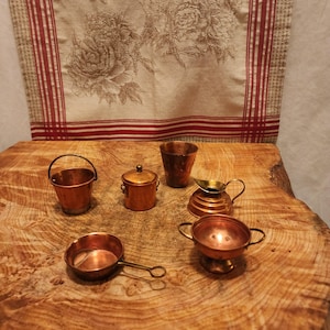 May include: A collection of miniature copper kitchenware, including a bucket, a pot with a lid, a cup, a small pitcher, a saucepan, and a bowl. The items are arranged on a wooden surface, with a floral patterned fabric in the background.