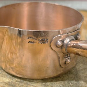 May include: A copper saucepan with a side handle, featuring a stamped mark that reads E. DEHILLERIN PARIS and MADE IN FRANCE. The metal surface is polished and reflective, showing a cylindrical body with a pouring spout and riveted handle attachment.
