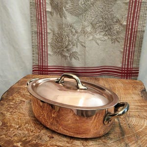 May include: An oval copper pot with a matching lid and two brass handles on the sides. The pot features a polished metallic finish and a central handle on the lid. The construction is seamless with a smooth, reflective surface throughout the exterior.