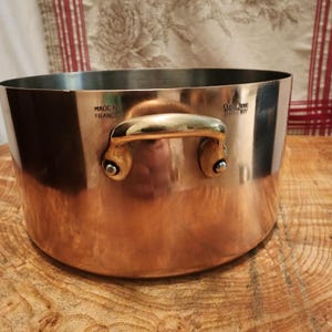 May include: A copper cooking pot with a brass handle and rivets. The pot has a shiny, reflective surface and a stainless steel interior. The words "MADE IN FRANCE" are visible. A vintage kitchenware item.