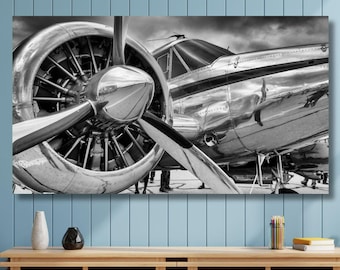 May include: A black and white wall art print featuring a close-up of a vintage propeller airplane. The image highlights the metallic sheen of the engine cowling, the large propeller blades, and the reflective fuselage against a cloudy sky background.