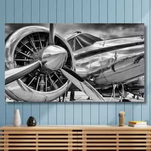 Könnte beinhalten: Schwarz-Weiß-Fotografie eines Oldtimer-Flugzeugs, die den Propeller und den Motor detailliert zeigt. Das polierte Metall reflektiert die Umgebung, mit Fokus auf dem vorderen Bereich des Flugzeugs. Das Bild hängt über einem Holzschrank.