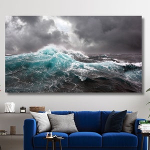Może przedstawiać: Duży wydruk na płótnie przedstawiający burzliwą scenę oceanu. Obraz przedstawia dramatyczny widok potężnych fal z turkusowymi grzywami rozbijającymi się o ciemne, wzburzone wody pod ponurym, szarym niebem. Grafika jest wyświetlana nad niebieską sofą.