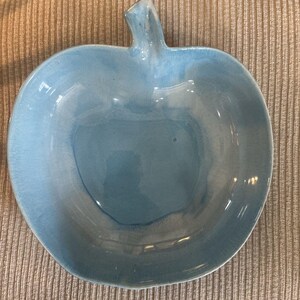 May include: A light blue ceramic apple-shaped bowl. The bowl has a stem and a glossy finish. The interior of the bowl is a lighter shade of blue than the exterior. The bowl is on a beige corduroy surface.