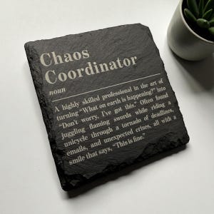May include: A black slate coaster with the words "Chaos Coordinator" engraved in white. The definition of a chaos coordinator is also included. A small potted plant is in the background.