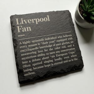 May include: A black slate coaster with the words "Liverpool Fan" and a definition. The definition describes an optimistic individual with encyclopedic knowledge of past glories. A small potted plant is in the background.
