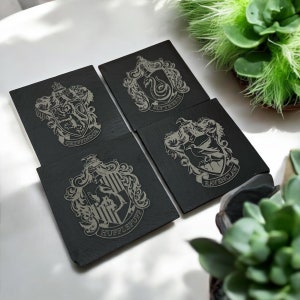 May include: A set of four black slate coasters, each engraved with a Hogwarts house crest: Gryffindor, Slytherin, Ravenclaw, and Hufflepuff. The designs are etched in a light grey colour.