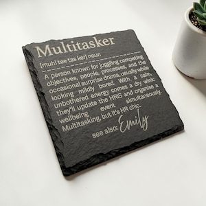May include: A dark grey slate coaster featuring the word "Multitasker" and its definition in white text. The coaster is personalized with the name "Emily". A small plant in a white pot is visible in the background.