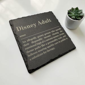 May include: A square black slate plaque with rough, uneven edges featuring white engraved text that reads: Disney Adult, noun, An allegedly grown person who spends their disposable income chasing the emotional high of a cartoon mouse.