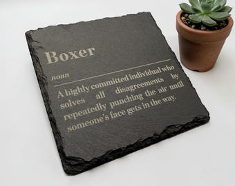 May include: A black slate coaster with the word "Boxer" and a definition etched in white. A small potted succulent sits to the right. The coaster's definition reads: "A highly committed individual who solves all disagreements by repeatedly punching the air until someone's face gets in the way."