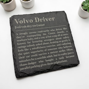 Op de afbeelding: Een vierkante zwarte leistenen onderzetter met de gegraveerde tekst Volvo Driver [vol-voh dry-ver] noun, gevolgd door een humoristische definitie die de rijgewoonten en persoonlijkheidskenmerken beschrijft die horen bij het besturen van een Volvo.