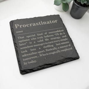 Könnte beinhalten: Ein quadratischer Untersetzer aus schwarzem Schiefer mit rauen, unebenen Kanten. Die Oberfläche ist mit dem Wort Procrastinator, der Bezeichnung noun und einer Definition graviert, die einen übermütigen Optimisten beschreibt.