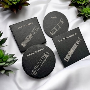 May include: Four black slate coasters with engraved lightsaber designs. Each coaster features a different Star Wars character: Darth Vader, Yoda, Obi Wan Kenobi, and Darth Sidious. The coasters are square and round.