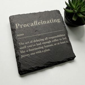 May include: A black slate coaster with the word "Procaffeinating" engraved in white. The definition of the word is also included. A small potted succulent is in the background.