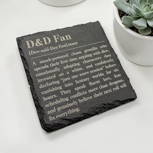 May include: A square black slate coaster with rough, uneven edges. The surface features white engraved text that reads: D&D Fan [Dee-and-Dee Fan] noun. A snack-powered chaos gremlin who spends their free time arguing with dice.