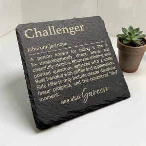Könnte beinhalten: Ein schwarzer Schieferuntersetzer mit dem Wort "Challenger" in weißer Schrift. Der Untersetzer enthält eine Definition des Wortes sowie die Worte "see also: Gareen". Im Hintergrund befindet sich eine kleine Topfpflanze.
