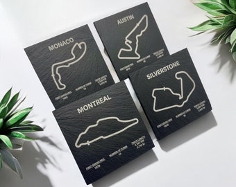 May include: Four black slate coasters with white engraved maps of famous race tracks. The coasters feature the names "Monaco", "Austin", "Montreal", and "Silverstone" along with the track length, number of turns, and first Grand Prix date.