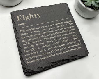 May include: A square black slate coaster featuring white engraved text that defines the word Eighty as a noun, describing the age with humorous phrases about joints, tea, naps, biscuits, and being a vintage model that is slightly creaky.