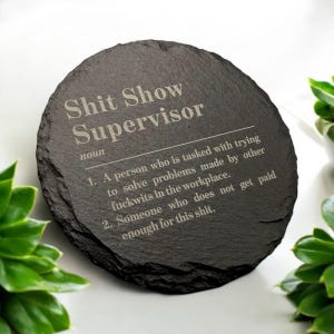 May include: A circular, dark grey slate coaster featuring the engraved phrase "Shit Show Supervisor" in a light grey font. The definition of the term is also engraved. Green plants surround the coaster.