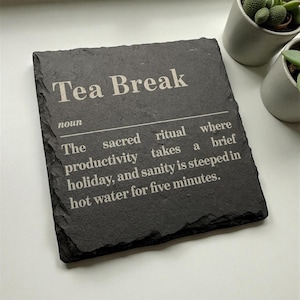 May include: A black slate coaster with the words "Tea Break" in white, and a definition. The definition describes a tea break as a sacred ritual. Two small potted succulents are in the background.