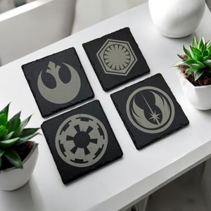 May include: A set of four square black slate coasters, each with a different grey graphic. The designs include a rebel alliance symbol, a first order symbol, an imperial symbol, and a rebel alliance starbird symbol.