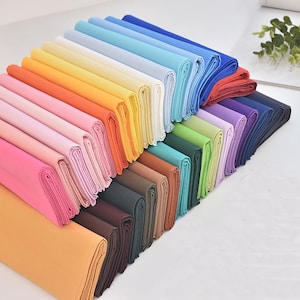 Può includere: Una collezione di tessuti piegati in una varietà di colori, tra cui rosa, arancione, giallo, verde, blu e marrone. Il tessuto è ordinatamente impilato, mostrando le diverse tonalità e trame. L'immagine è ben illuminata, evidenziando i colori vivaci dei tessuti.